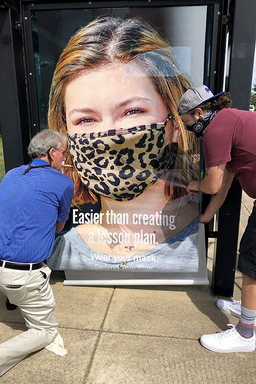 Ed Arning, left, MTSU director of market development, and senior advertising major Andrew Felts of Murfreesboro adjust a “Defend True Blue” promotional poster featuring senior advertising major Bella Utley of Lebanon, Tennessee, on a Raider Xpress bus shelter sign on campus. Felts and Utley are part of an MTSU School of Journalism and Strategic Media student team who created a public relations and advertising campaign to encourage their fellow students to wear their masks on campus. The campaign slogan, "Wear Your Mask. Defend True Blue," is part of each image. (MTSU photo)