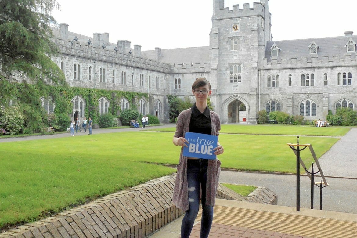 Angele Latham, a sophomore Visual Communications student, stands in front of a stone building on the Cork, Ireland campus holding a blue sign that reads 