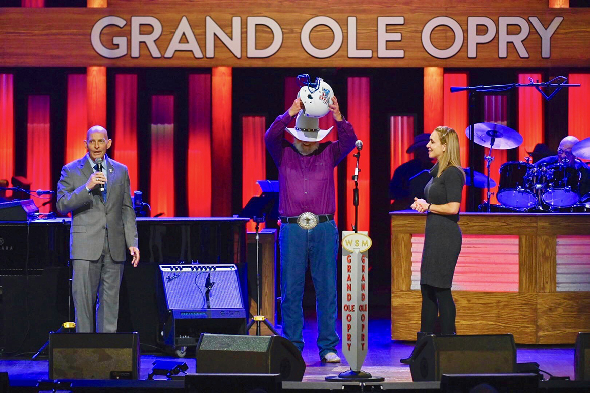 Retired Army Lt. Gen. Keith Huber, left, speaks to the audience at the Grand Ole Opry in Nashville, Tenn., after presenting Country Music Hall of Famer Charlie Daniels, namesake and patron of MTSU’s Charlie and Hazel Daniels Veterans and Military Family Center, with a Blue Raider Football helmet. Huber and Daniels Center Director Hilary Miller, right, surprised Daniels during his Oct. 15 show. (MTSU photo by Cat Curtis Murphy)