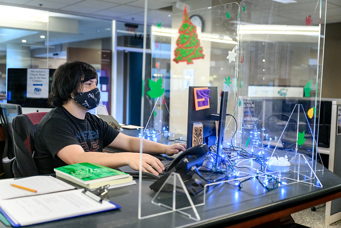 Sean Strickland, a student worker at Makerspace, staffs the help desk behind Plexiglass shields while wearing a mask to adhere to pandemic protocols. Figurines made with Makerspace equipment adorn the shields. (MTSU Photo by J. Intintoli)