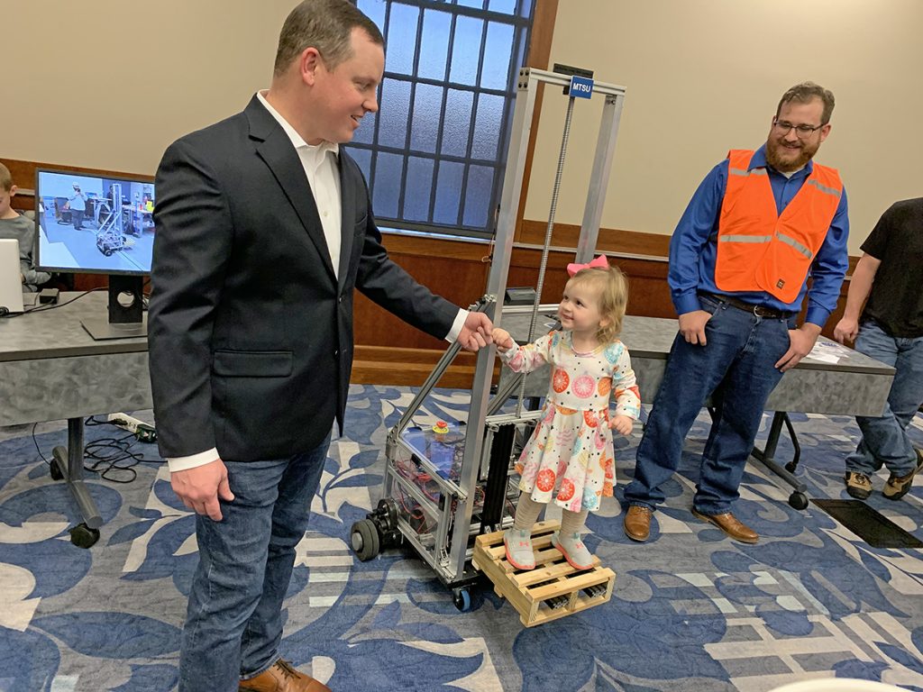 MTSU mechatronics engineering student J.R. Jackson, left, of Lebanon, Tenn., holds 3-year-old daughter Anna Kate’s hand as she rides up and down on the automated forklift his team designed and built for the recent Engineering and Technology Mech-Tech Expo. (MTSU photo by Randy Weiler)