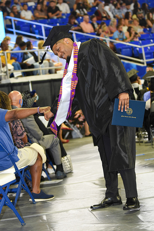A new MTSU Class of 2021 graduate bumps fists with a supporter as he makes his way back to his seat in Murphy Center, degree in hand, at the August commencement ceremony. He's wearing a white stole denoting his new degree in professional sales from the Jones College of Business and the purple-and-gold Kente stole of his fraternity, Omega Psi Phi. MTSU students graduating Saturday, Dec. 11, in the fall 2021 commencement can purchase and wear special new stoles from the university's Phillips Bookstore representing their colleges. (MTSU file photo by GradImages)