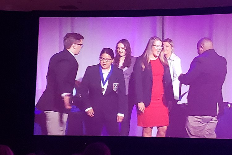 MTSU Collegiate DECA students Allison Mullins, an MBA major, second from left, and marketing major Olivia Andersen, third from right, are shown on a video screen receiving their medals for making it to the final round of the event planning team event at the 2022 DECA’s Career Development Conference being April 9-12 in Baltimore, Md. (Submitted photo)