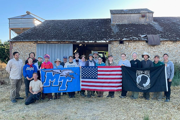 Middle Tennessee State University students and faculty involved with MTSU’s Forensic Aviation Archaeology study abroad course, along with international volunteers, display an MTSU banner and an American flag near the site of their grant-funded field recovery project in France where students help search for remains of missing U.S. World War II personnel in August 2022. (Photo courtesy of Tiffany Saul)