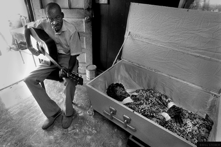 With guitar in hand, legendary blues musician and folk artist Son Thomas sits in the front room of his Leland, Miss., home next to a sculpted depiction of a woman in a casket, made from clay. The circa YEAR? image is one of many on display as part of the Middle Tennessee State University Baldwin Photographic Gallery exhibit, "Deep Roots: Evocations of the Mississippi Blues," featuring photos by alumnus Bill Steber, on display through Dec. 9, 2023. (Photo courtesy of Bill Steber)