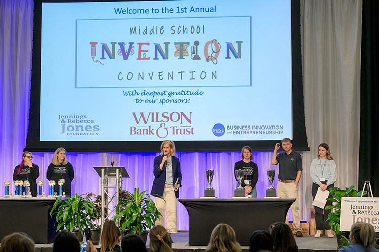 Middle Tennessee State University professor Tracey Huddleston, center, speaks to the crowd of over 250 at the inaugural Middle School program of the annual Invention Convention in the Student Union Ballroom on Feb. 22, 2024. Sharing the stage with Huddleston, from left, are master’s education students Carrie Matherly, Sarah Lutz, Huddleston, master’s education student Estella Pennell, and education faculty Jeremy Winters and Katie Schrodt. The College of Education puts on the annual Invention Convention to host local students to showcase their range of creative, innovative inventions. (MTSU photo by Andy Heidt)