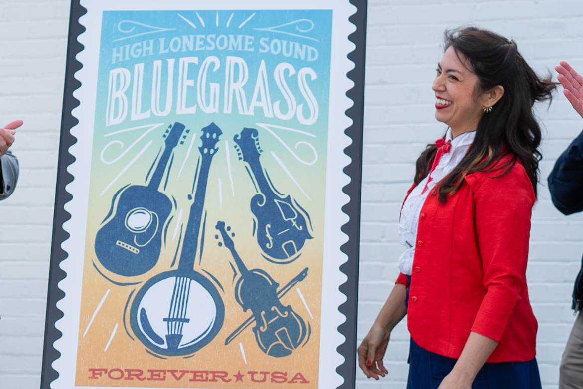 Middle Tennessee State University graphic design alumna Heather Moulder stands next to a large version of the High Lonesome Sound Bluegrass forever stamp that she designed for the United State Postal Service at an unveiling ceremony at a post office in Readyville, Tenn., in April. (Photo courtesy of Daxton Patrick)