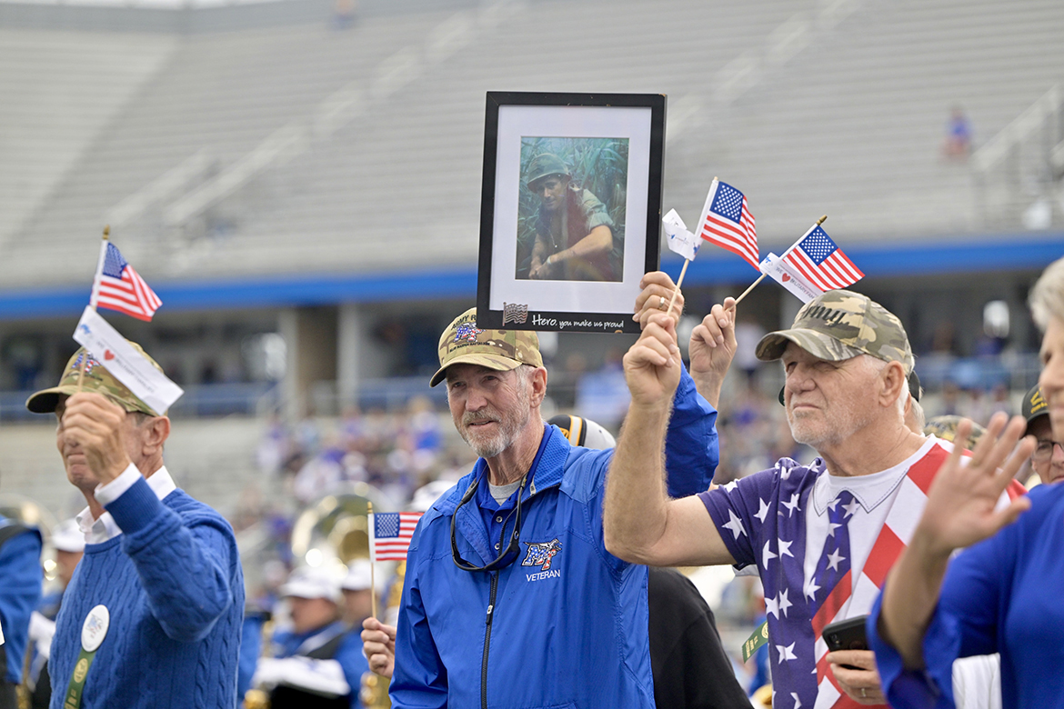 Middle Tennessee State University alumnus and U.S. Army Veteran Bob Lamb (Classes of 1969 and ’77), center, of Murfreesboro, Tenn., walks across Horace Jones Field in Floyd Stadium in Murfreesboro carrying a framed photo of his longtime friend and veteran, Bud Morris (’69, ’75),  in November 2024 during the 42nd annual MTSU Salute to Veterans and Armed Forces game during the halftime parade by service branches to theme songs performed by the Band of Blue. Morris, who attended the game, saluted his friends and fellow vets from the Jeff Hendrix Stadium Club, the first time in 40 years he has not walked with his military brothers and sisters. (MTSU file photo by Andy Heidt)