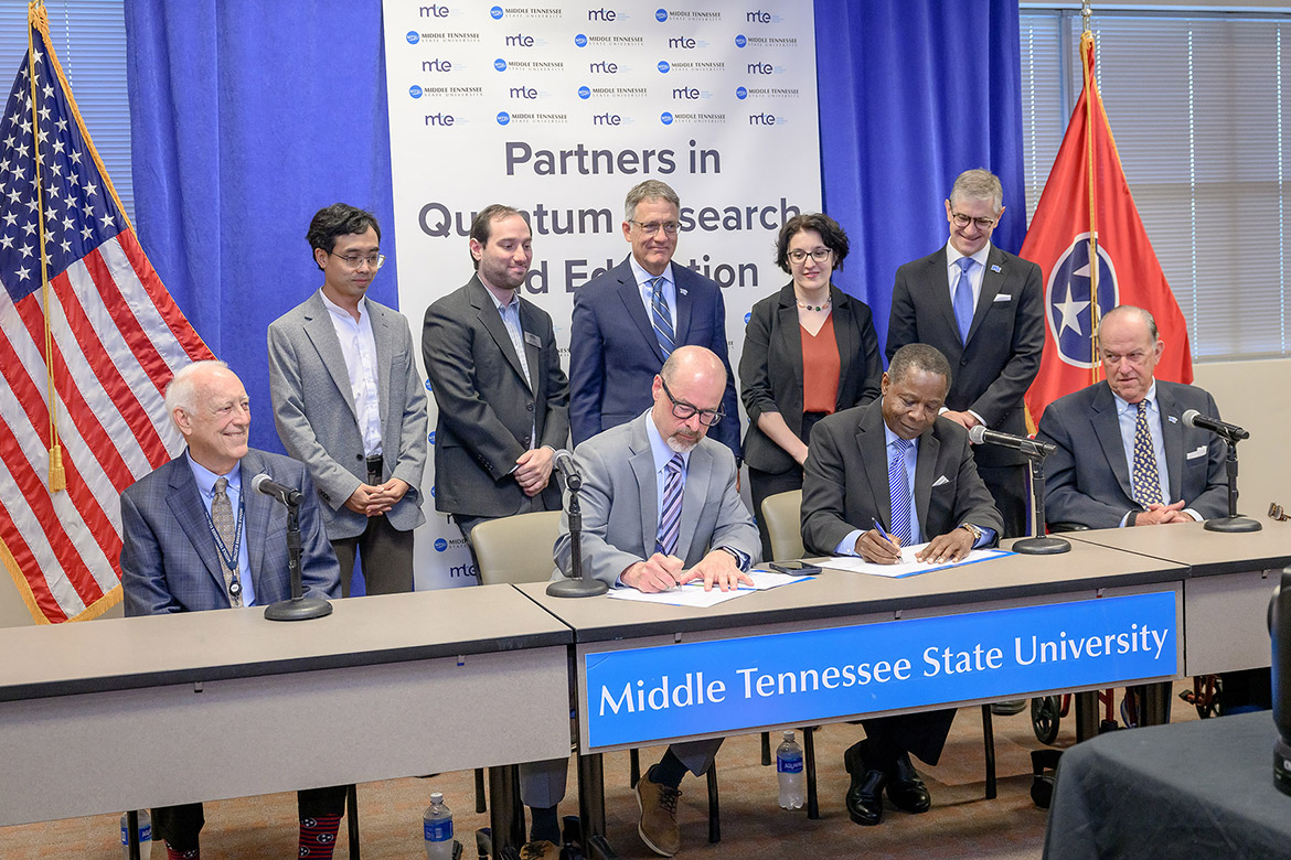 Two men seated at table signing documents with several people looking on behind and beside them.