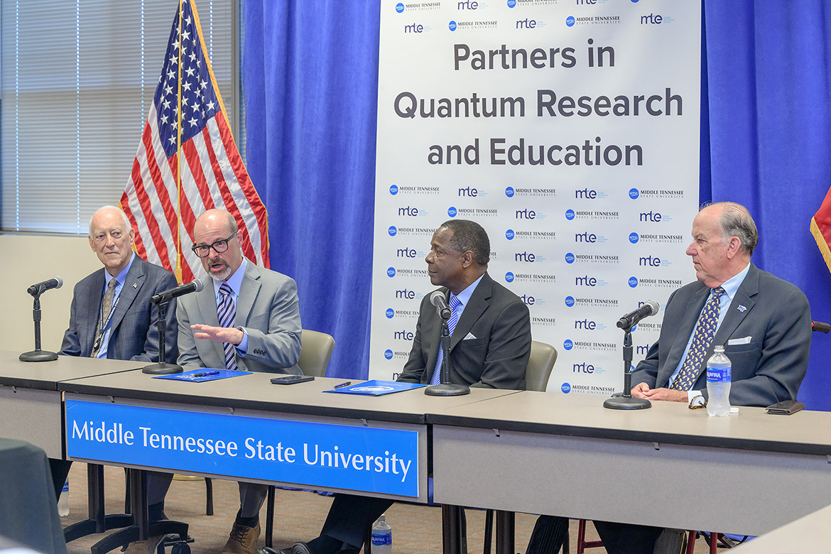 Four men sitting at table with microphones in front of them, with one of them speaking.