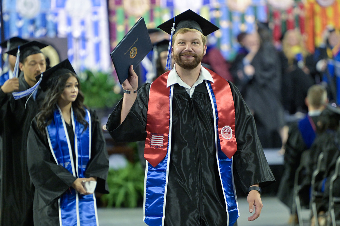 Wearing the special red stole designating his status as a student veteran, a proud Class of 2026 graduate holds up his diploma cover.