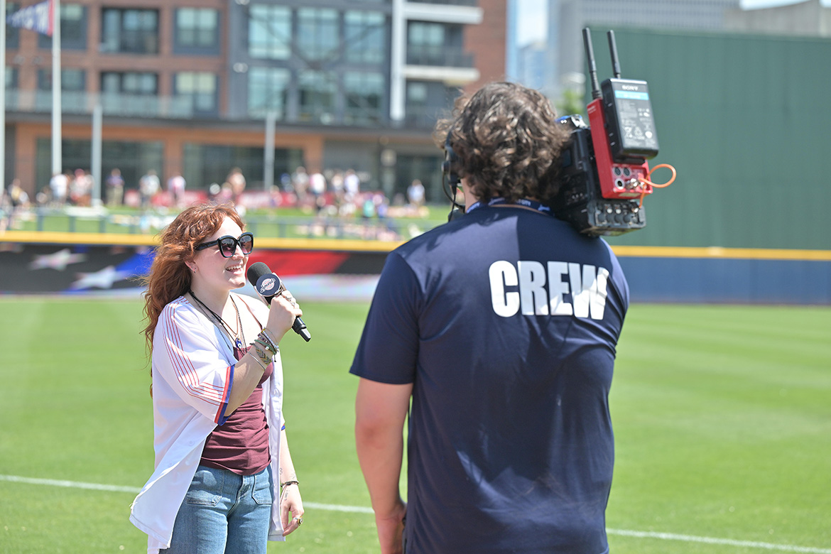 Woman singing national anthem on baseball field with camera operator in the foreground.
