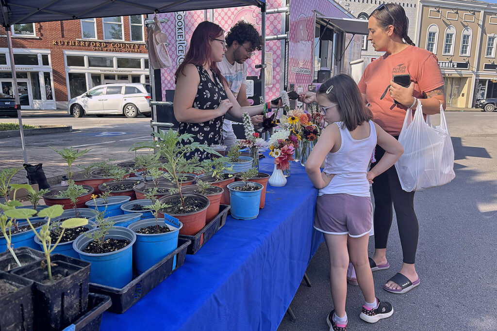 Soil science majors from Middle Tennessee State University in Murfreesboro, Tenn., Alyssa Sparr, left, of Bradford, Tenn., and James Terrell from Franklin, Tenn., staff the MTSU Small Farm booth on June 6, 2026, at the Main Street Murfreesboro Saturday Market, as Murfreesboro mom Monica Bycroft, far right, and her daughters Norah and Maeve, finalize their purchase. The Saturday Market is held in downtown Murfreesboro from 8 a.m. to noon each Saturday from May through October. (MTSU photo by Nancy DeGennaro)