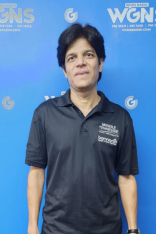 Sanjay Asthana, journalism professor, standing in front of a blue backdrop at the WGNS Radio studio.