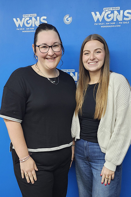 MTSU Jones College of Business students Savanah Gill and Ella Cobb standing in front of blue backdrop the inside WGNS Radio studio.