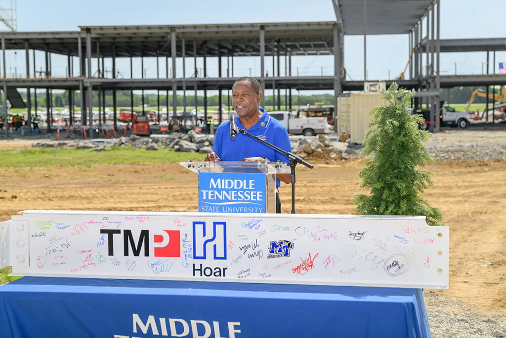 Sidney A. McPhee, president of Middle Tennessee State University in Murfreesboro, Tenn., gives remarks at the topping-out ceremony held Tuesday, July 21, 2026, for the MTSU Aerospace Department’s $62 million flight training center, under construction in the background, at the Shelbyville Municipal Airport in Bedford County. In front of McPhee is the ceremonial beam that was later placed at the highest point of the structure. (MTSU photo by J. Intintoli)