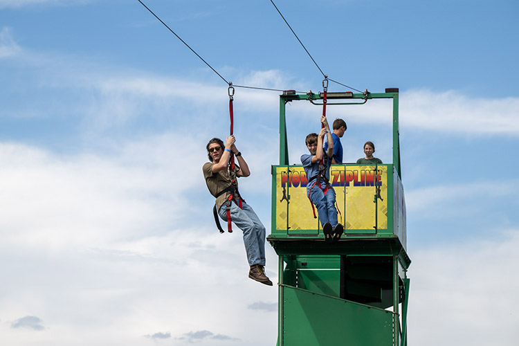Two students dangle from a zipline over the Student Union Commons against blue skies.