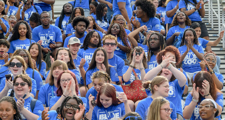 Students wearing blue shirts clap and cheer from stadium seats.