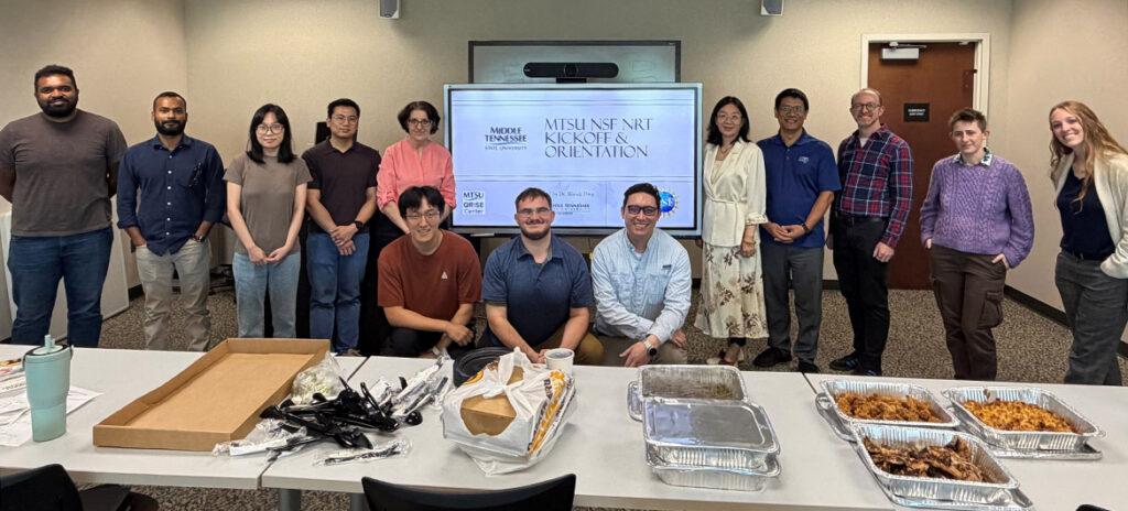 A group of 13 MTSU students and faculty pose in a line in front of a table of food. 