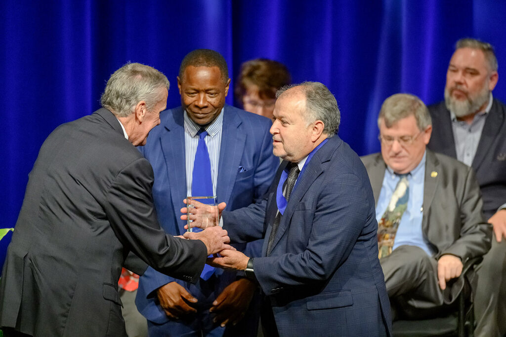 Lalo Davila, professor of music and director of percussion studies at Middle Tennessee State University in Murfreesboro, Tenn., accepts the 2026 Career Achievement Award from Mike Ussery, vice president of the MTSU Foundation, at left, as President Sidney A. McPhee, center, and other top administrators, seated, watch the presentation at the annual Fall Faculty and Staff Meeting held Thursday, Aug. 20, in Tucker Theatre on campus in Murfreesboro, Tenn. Davila, an award-winning percussionist and educator, has spent the past four decades at MTSU in the School of Music. (MTSU photo by Andy Heidt)