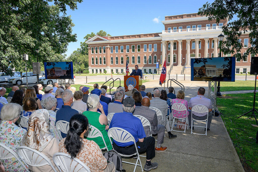 Computer science graduate student Gavin Liles stands behind a podium in front of Kirksey Old Main and speaks to the audience about the building's significance. 