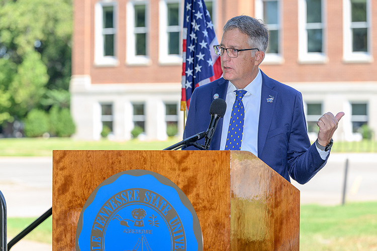 MTSU Provost Mark Byrnes stands behind a podium, gesturing towards the newly remodeled Kirksey Old Main building. 