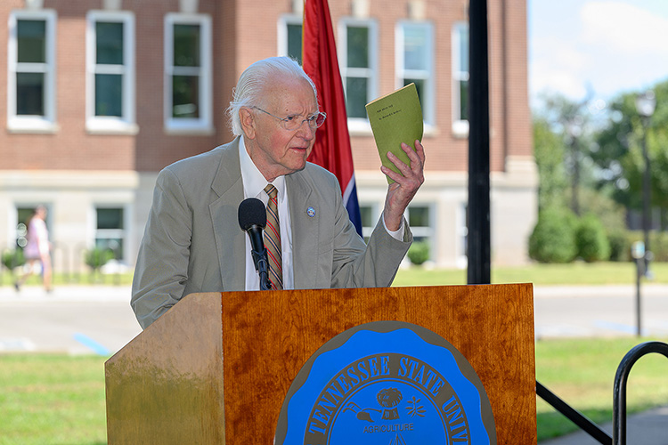 Howard Graden Kirksey Jr. stands behind a podium and holds up a small green booklet.