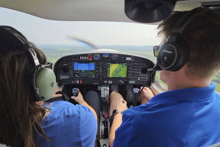 This photo shows two people from behind, both wearing blue shirts, sitting side-by-side in the cockpit of a small plane. The view through the windshield shows rolling green hills.