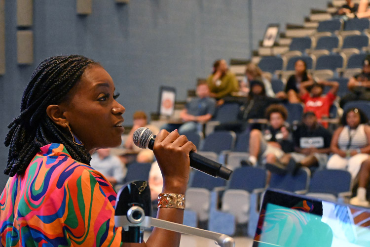 A woman with dark skin and braids wearing a colorful dress speaks into a microphone as an auditorium full of students sitting in blue chairs listen.