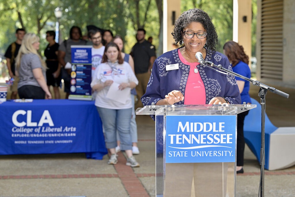 A woman reading a document standing behind a lectern outside.