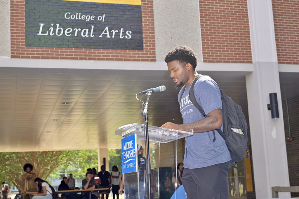 A male college student reading a document standing behind a lectern outside.