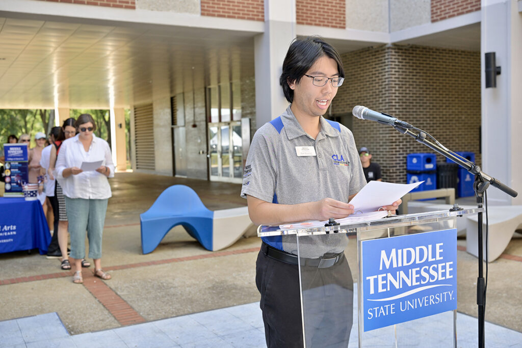A man reading a document standing behind a lectern outside.