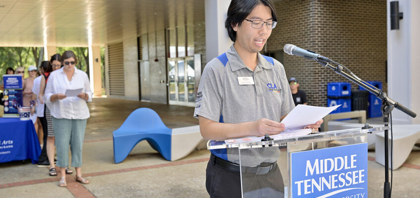 A man reading a document standing behind a lectern outside.