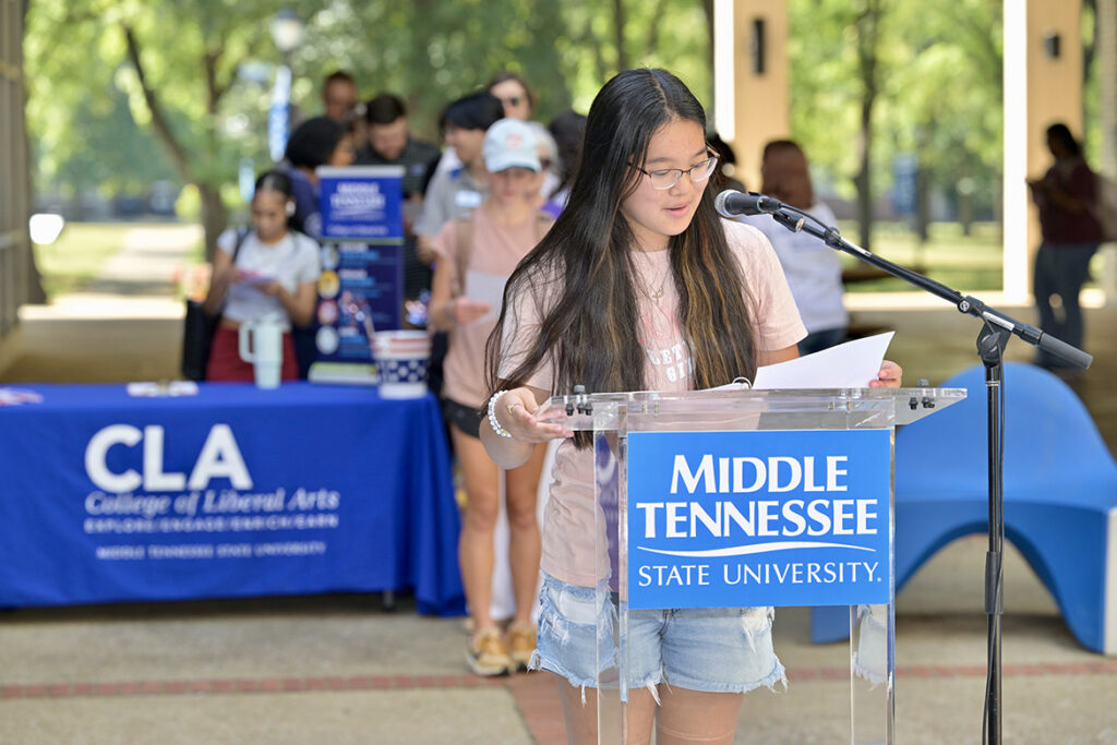 A female college student reading a document standing behind a lectern outside.