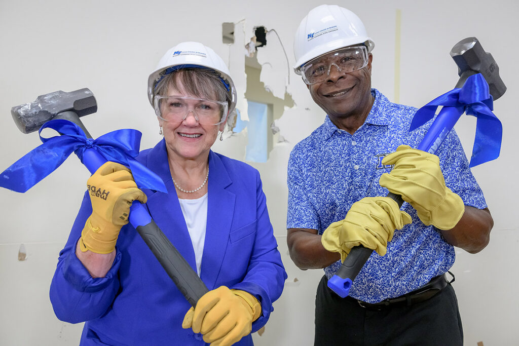 A woman and man smiling, wearing hard hats, safety goggles and gloves holding sledgehammers.