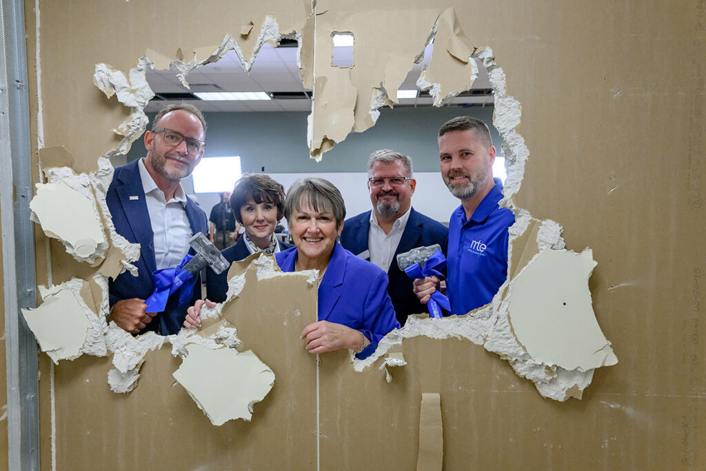 A group of five people standing and smiling, looking through a large hole in a wall.