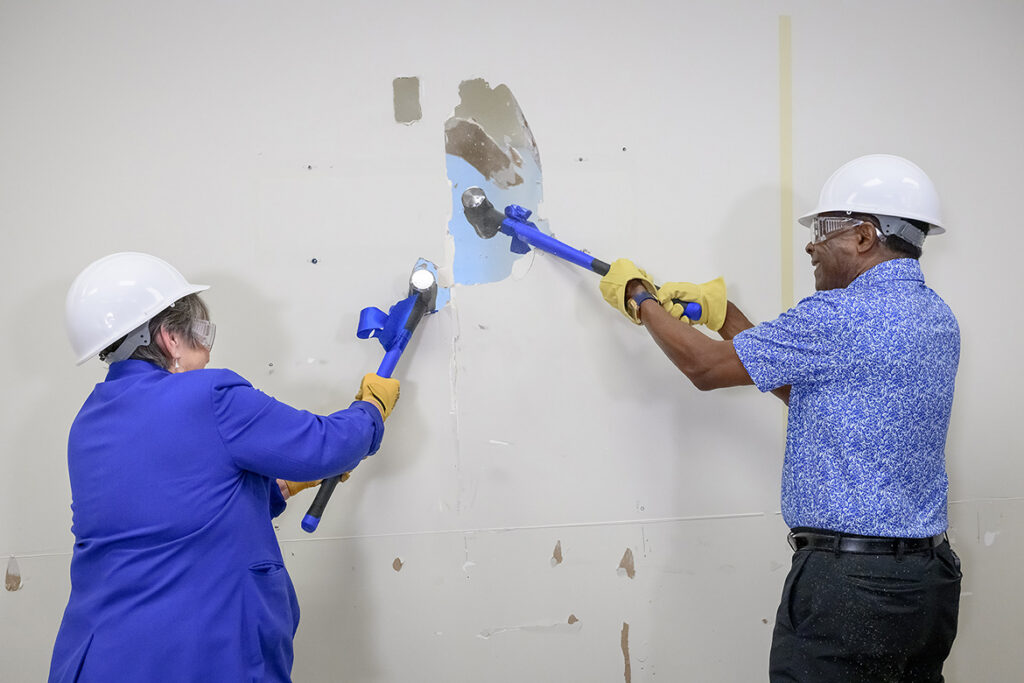 Woman and man swinging sledgehammers to knock holes in a wall.