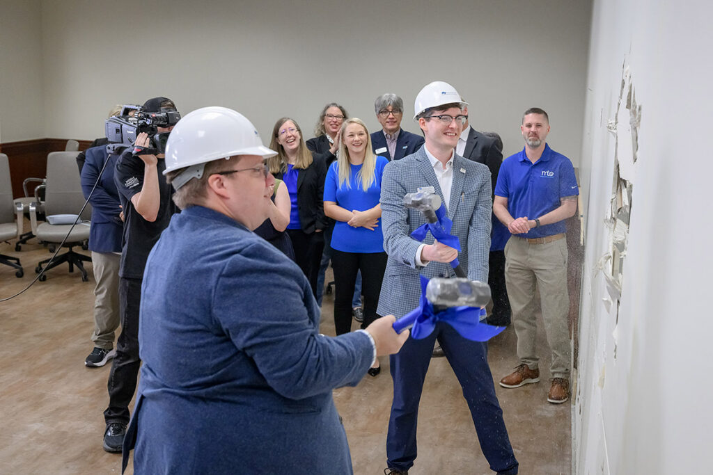 Two men holding sledgehammers preparing to strike a wall as several people look on in the background.
