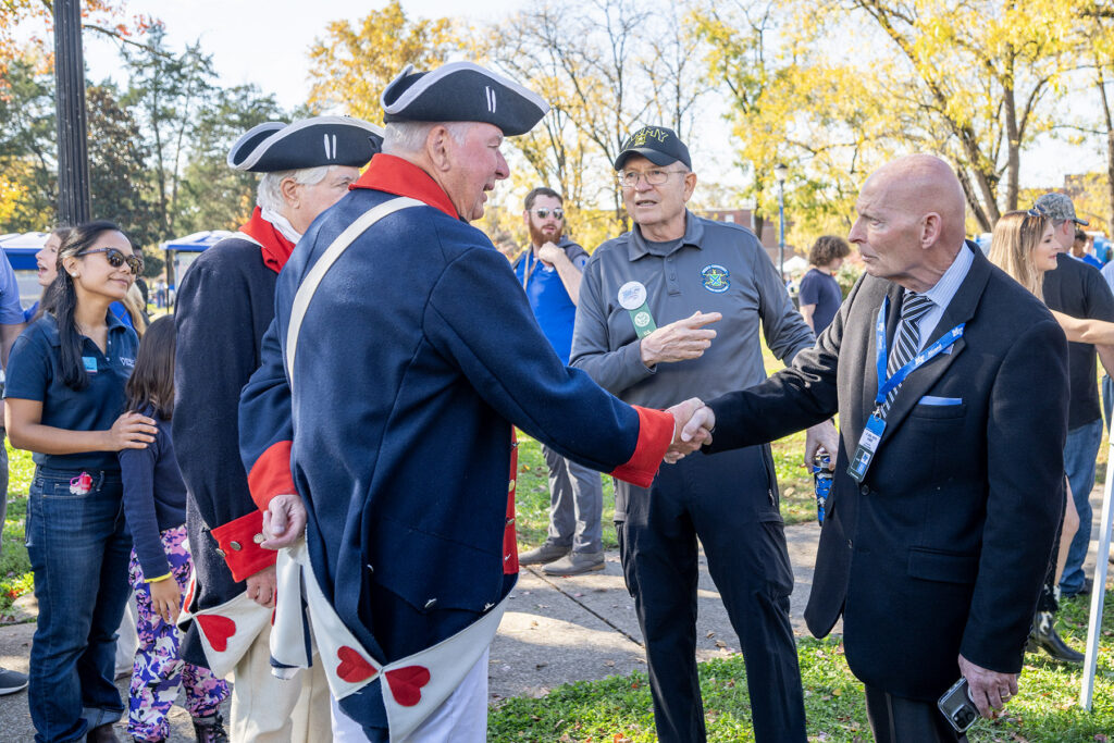 A bald man in a dark suit shakes hands with a man wearing a tricorn hat.