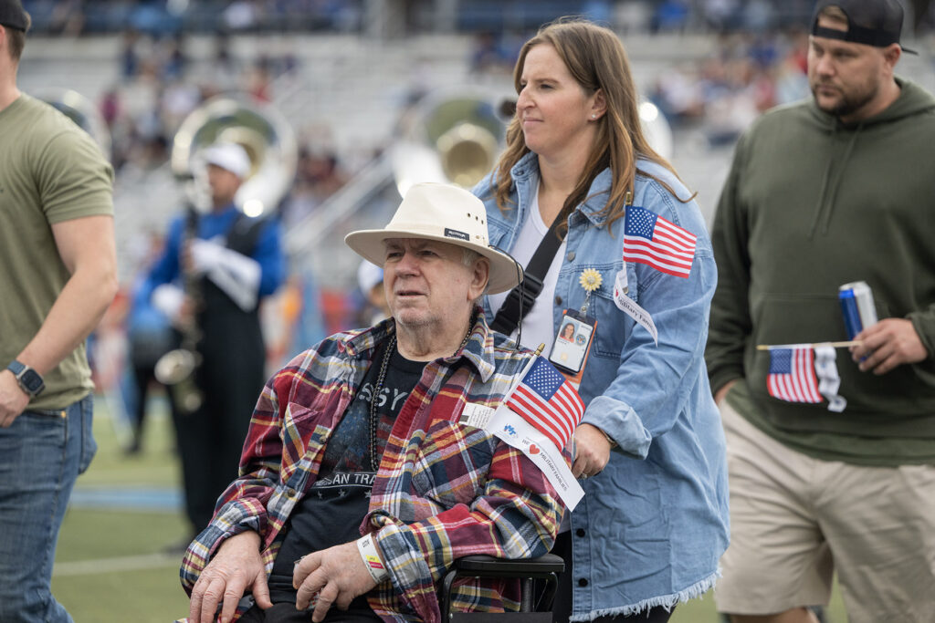 A woman in a blue denim shirt pushes an older man in a plaid shirt and hat in a wheelchair, both holding American flags.