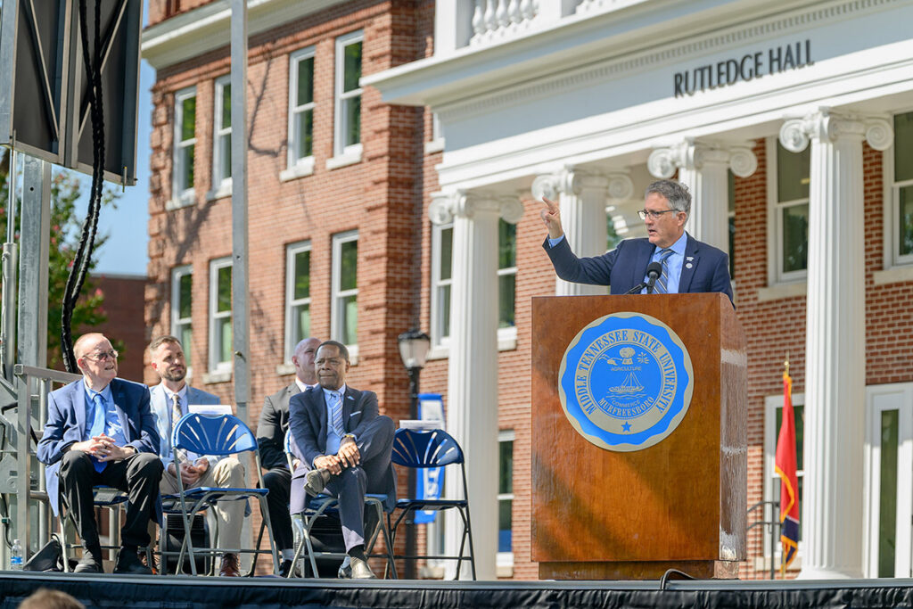 A man in a dark blue suit and tie stands behind a podium, pointing towards the audience. 