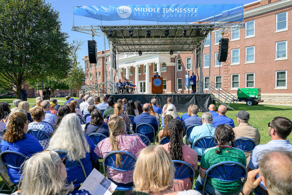 A crowd of people seated in blue folding chairs in front of a atage, where a man stands behind a podium. 