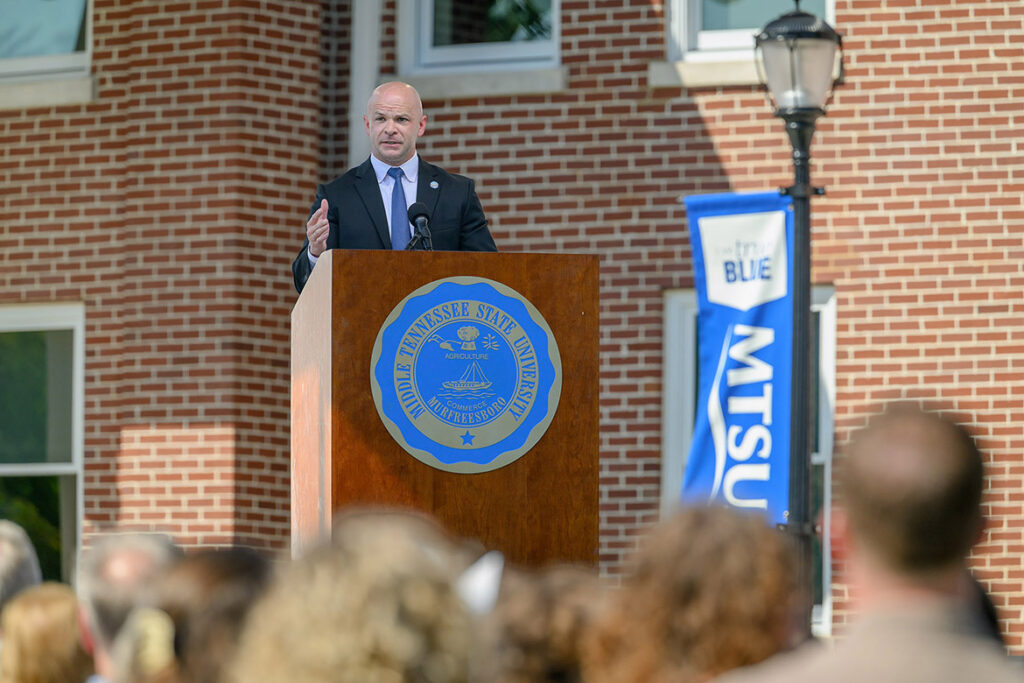 A man in a gray suit and blue tie stands behind a podium. 