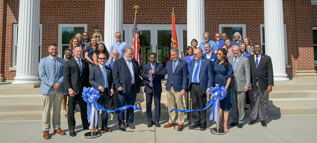 A group of people in suits and nice cloths stand on the front steps of a building. One man holds a giant pair of scissors with a blue handle.
