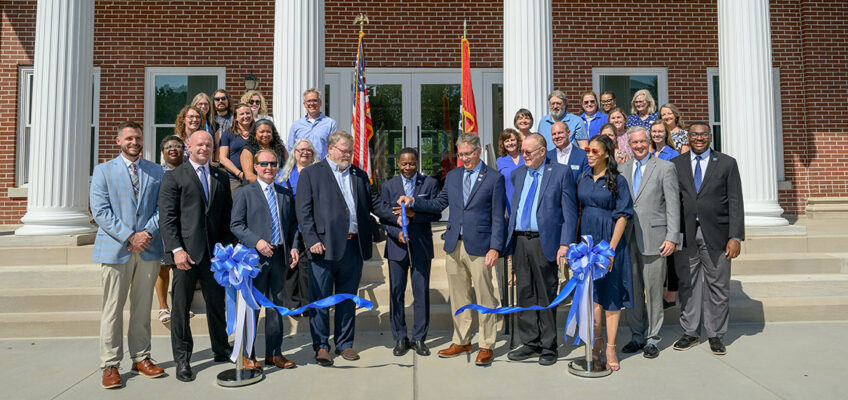 A group of people in suits and nice cloths stand on the front steps of a building. One man holds a giant pair of scissors with a blue handle.