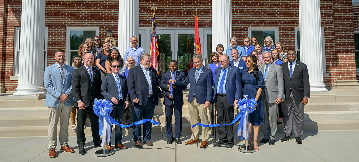 A group of people in suits and nice cloths stand on the front steps of a building. One man holds a giant pair of scissors with a blue handle.