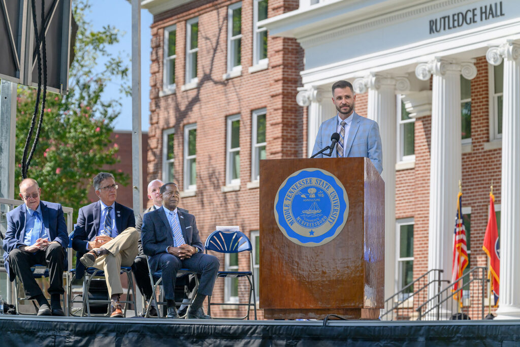 A bearded man in a light blue plaid suit and tie stands behind a podium. 