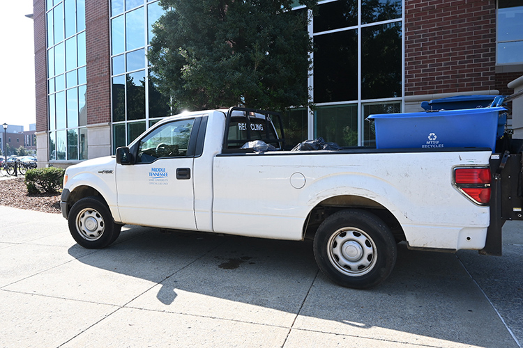 White pickup truck with blue recycling bins in back parked outside library building.
