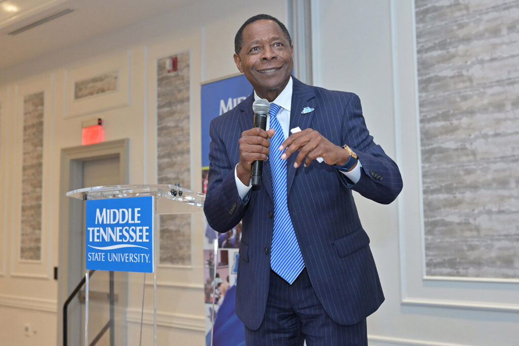 A man in a dark blue pinstriped suit smiles, holding a microphone and standing beside a podium.