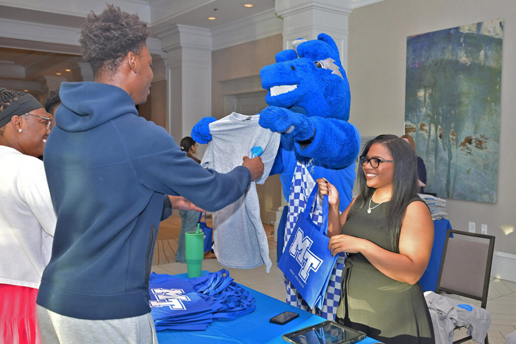 A person in a giant blue horse costume hands a tshirt to a high school student.
